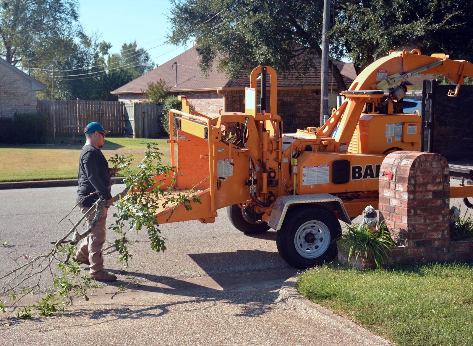 Worker feeding a cut tree branch into an orange wood chipper on a residential street during a neighborhood cleanup
