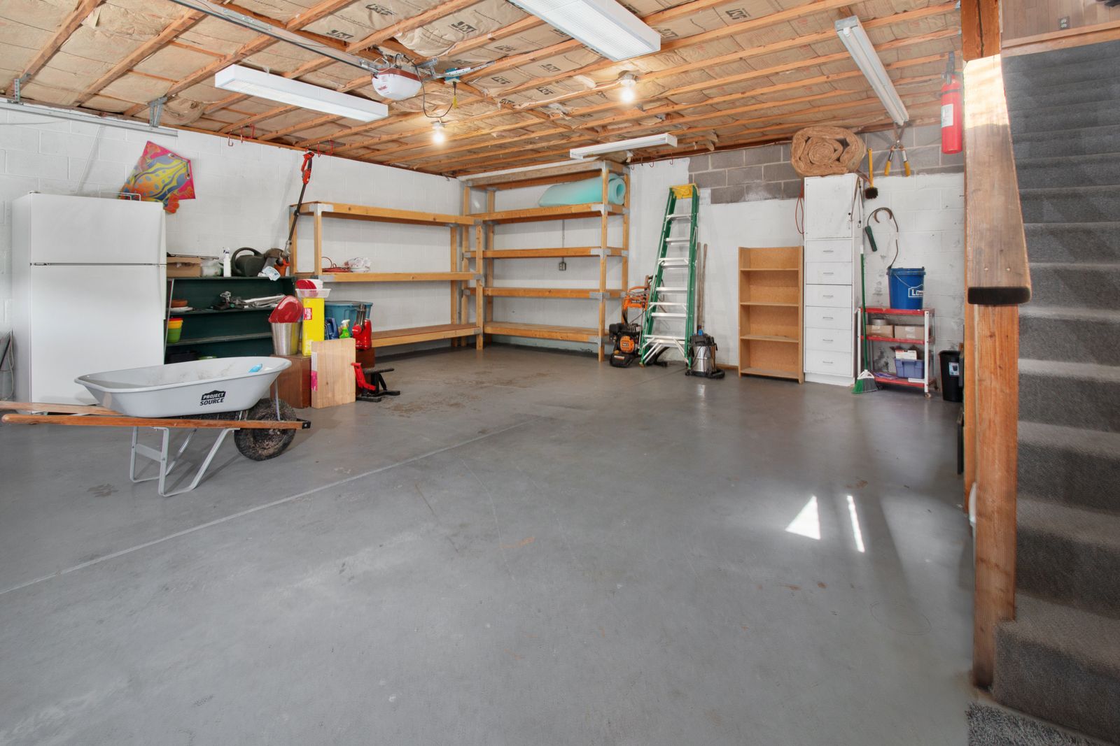 Inside a home garage with storage shelves, tools, a ladder, a wheelbarrow, and a ceiling-mounted garage door opener