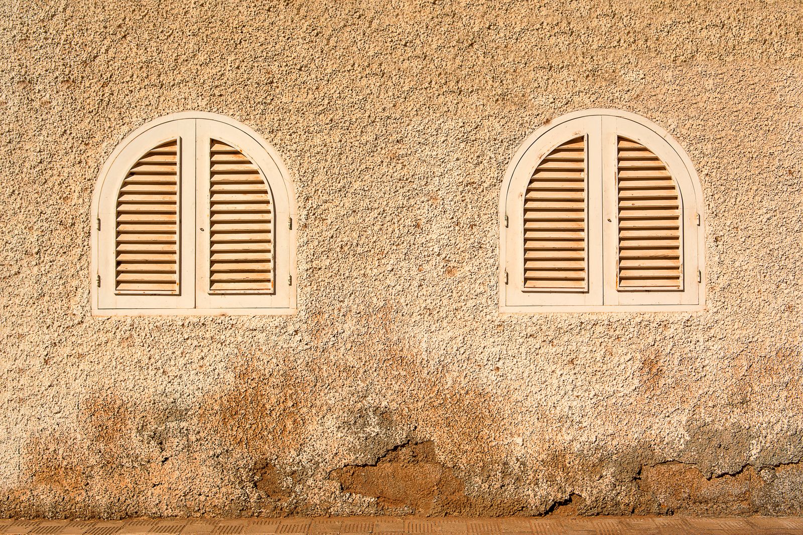 Two arched windows with closed louvered shutters on a stucco wall